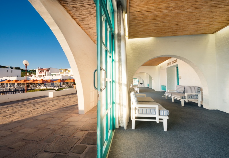 Shaded white-arched veranda with turquoise doors, striped cushioned lounges under bamboo ceiling overlooking waterfront terrace with orange umbrellas.