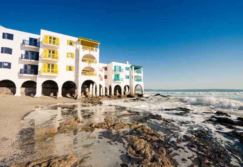 Seafront hotel on arched stilts with bright yellow and teal balconies, rocky beach and crashing surf.