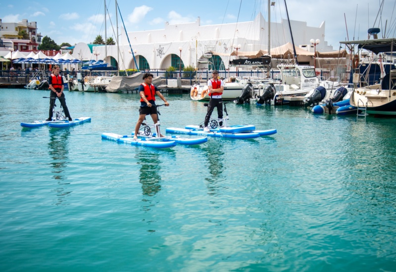 Three guests in red life vests riding blue water-bikes in a turquoise marina with moored yachts and white seaside buildings.