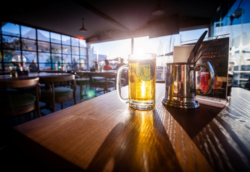 Sunlit beer mug on a wooden table in a waterfront restaurant, metal cutlery holder and menu, warm relaxed atmosphere.