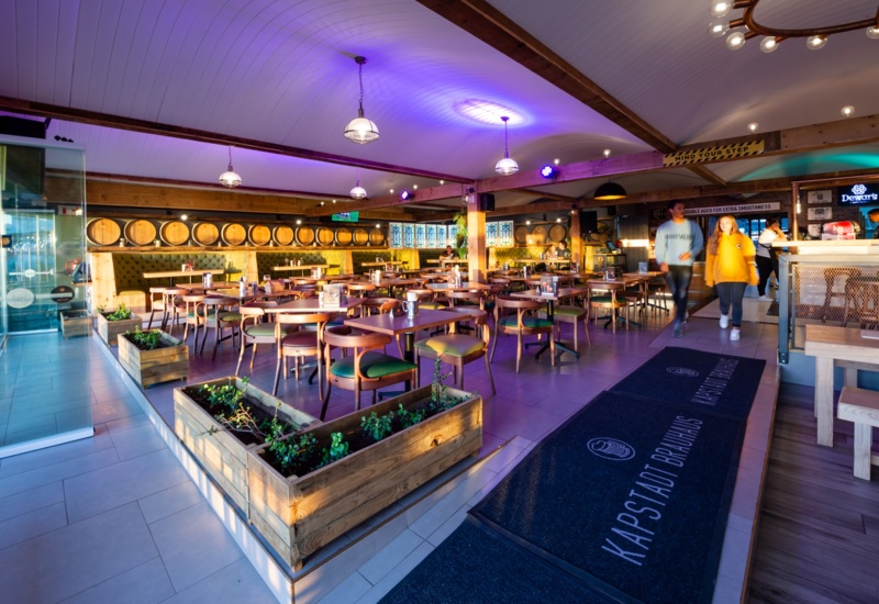 Brewery-style dining area with barrel-lined wall, wooden tables, planter boxes, purple lighting and guests at the bar.