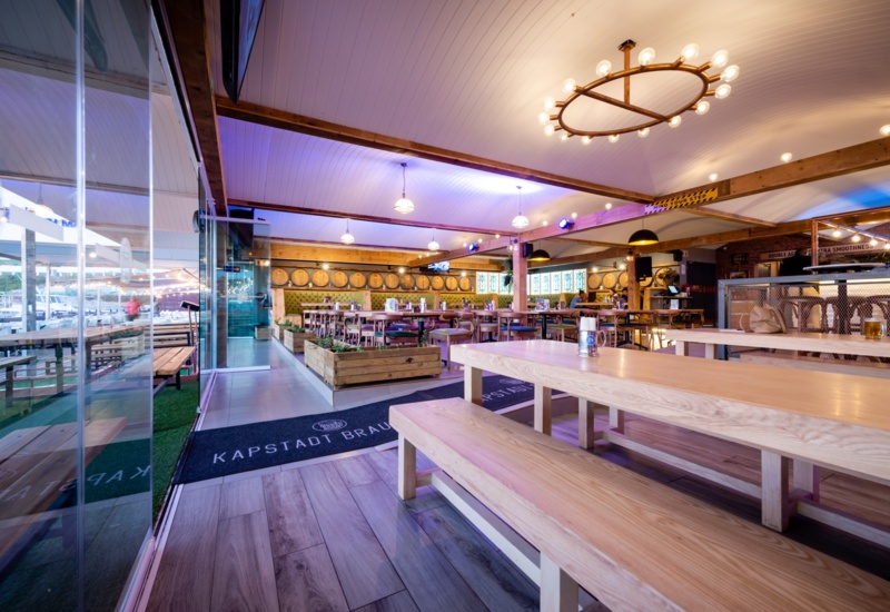 Waterfront restaurant with long communal wooden tables and benches, barrel-lined wall, warm lights and marina views.