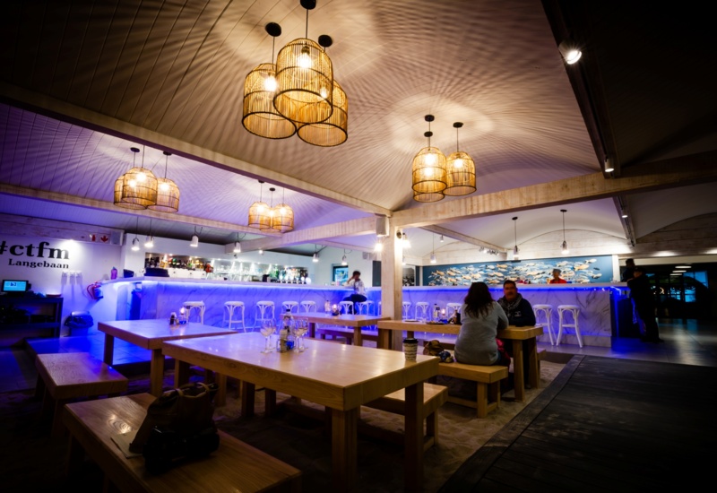 Casual beachfront bar and restaurant with communal wooden tables and benches, blue-lit marble bar, rattan pendant lights, couple dining, fish wall art.