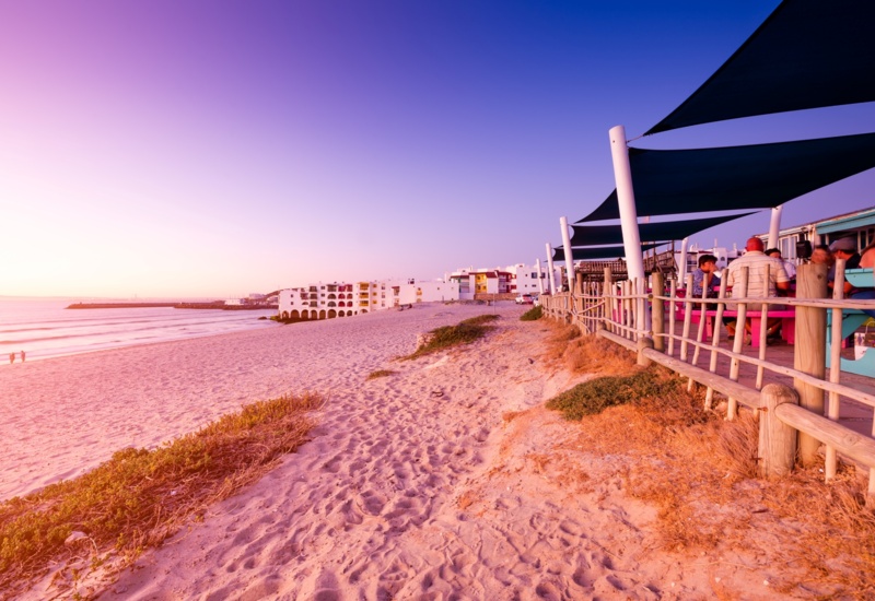 Beachfront restaurant patio with shaded seating and diners beside a sandy path leading to white arch-fronted seaside hotel at sunset.