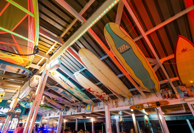 Beachfront bar interior with surfboards and sails suspended from exposed timber beams, colorful LEDs, lively dining.