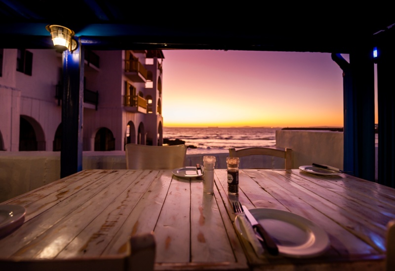 Covered beachfront terrace with rustic wooden table set for four, salt-and-pepper shakers, arched hotel balconies and a vivid sunset over the ocean