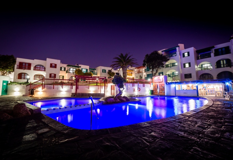 Moonlit pool glowing electric blue, set amid white Mediterranean rooms with balconies, palm tree and poolside bar.