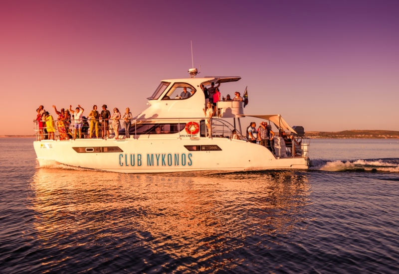 Club Mykonos party yacht with guests mingling on open decks at sunset, panoramic coastal views and warm purple-pink sky.