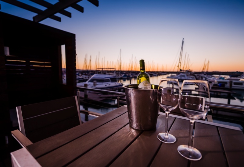 Waterfront terrace table with chilled bottle in an ice bucket and two wine glasses, overlooking yachts at sunset in a marina.