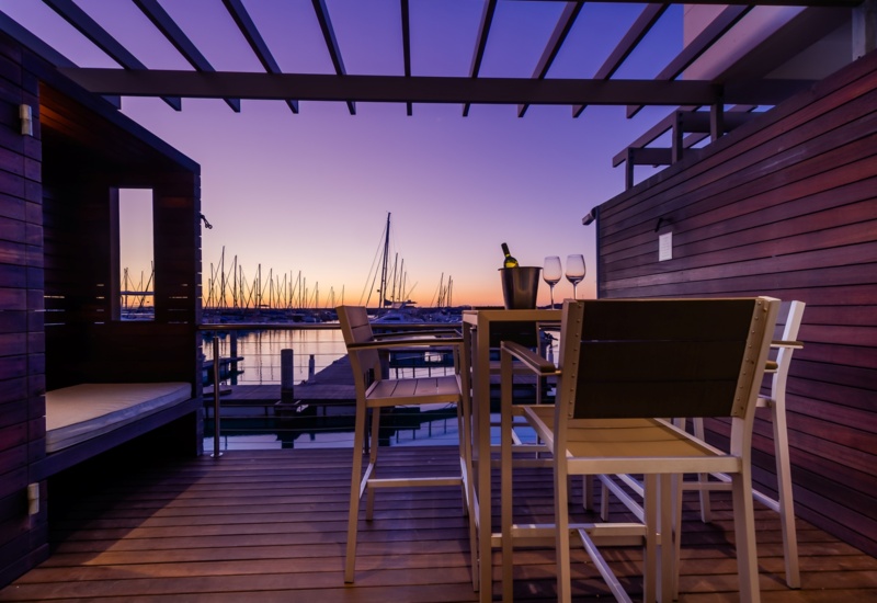 Private wooden deck with high table, wine bottle and glasses, cabana bench overlooking marina and sailboats at sunset.