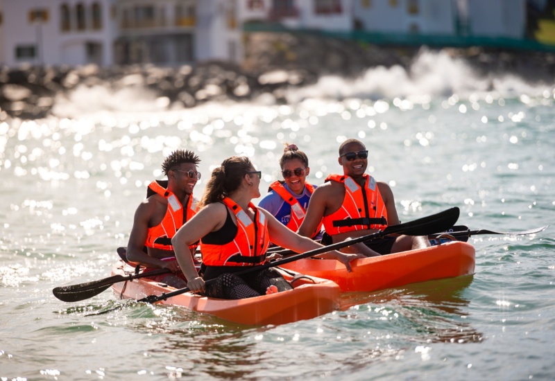 Four guests in orange life vests kayaking bright orange kayaks on sunlit, sparkling sea with resort-lined coastline