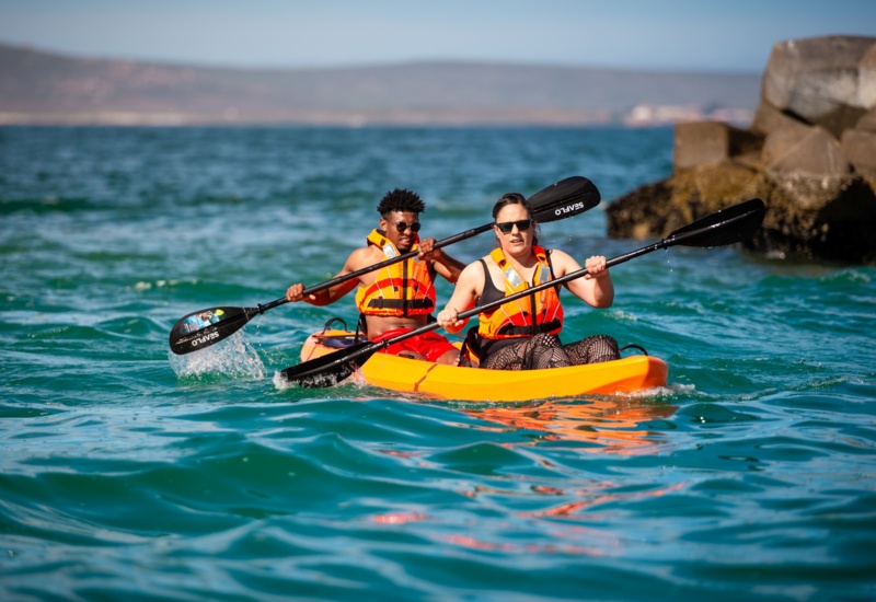 Two guests in an orange tandem kayak paddling turquoise water near rocky shore, both wearing life jackets.
