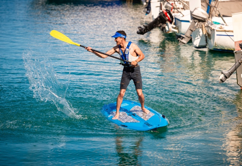 Guest paddleboarding on a blue board at a marina, wearing life vest and visor and splashing water beside docked boats
