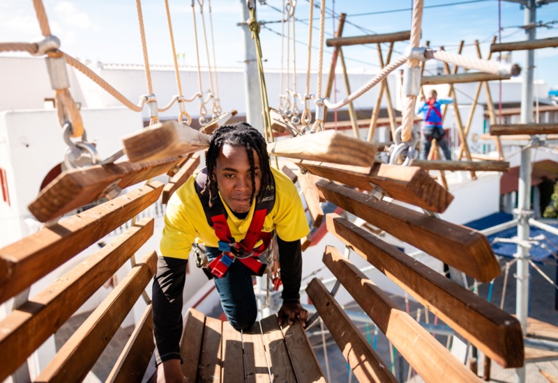 Guest in safety harness crawling through suspended wooden ropes-course planks on an outdoor adventure course at a sunny resort