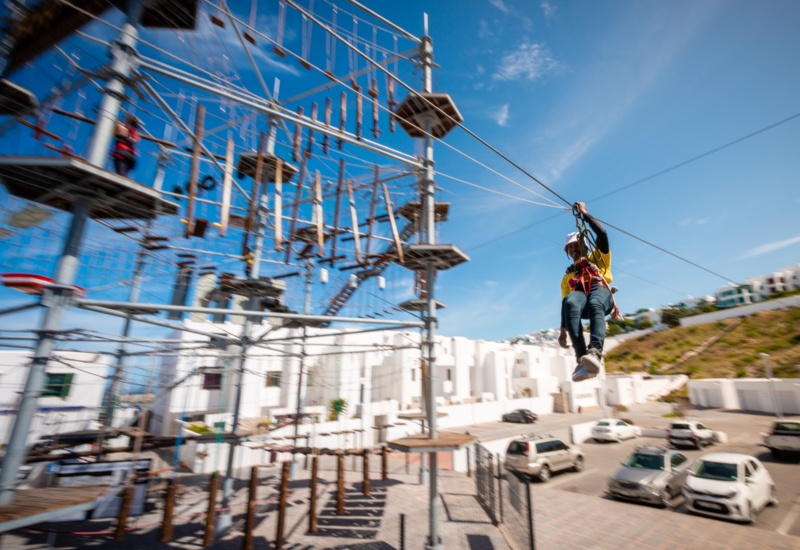 Guest wearing helmet and harness ziplines from elevated ropes course above white resort buildings and parking lot on a sunny day.