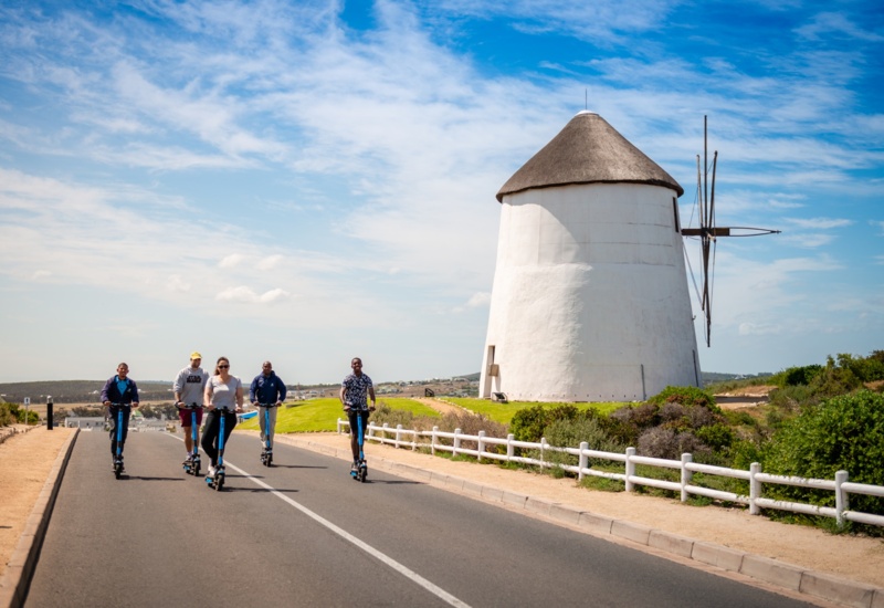 Five people riding electric scooters along a sunny coastal road past a white thatched windmill and low scrub