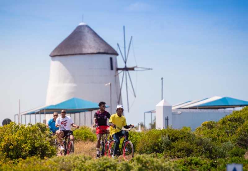 Guests cycling past a white coastal windmill and blue-canopied pavilions through sunlit coastal scrubland