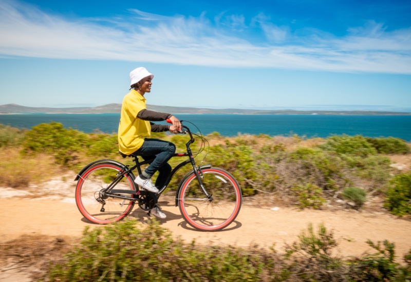 Guest cycling a rental bike along a sunlit coastal trail, turquoise ocean backdrop, relaxed casual attire and white bucket hat