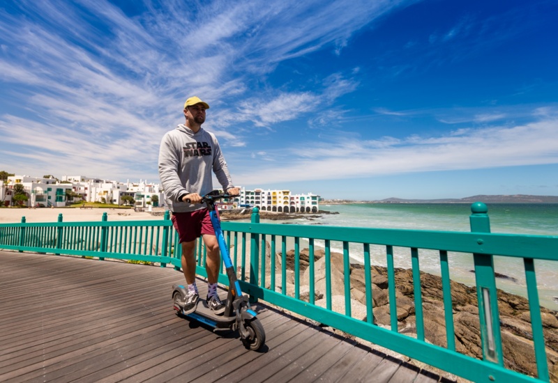 Man riding an electric scooter along a sunny seaside boardwalk with turquoise railing, ocean views and beachfront apartments