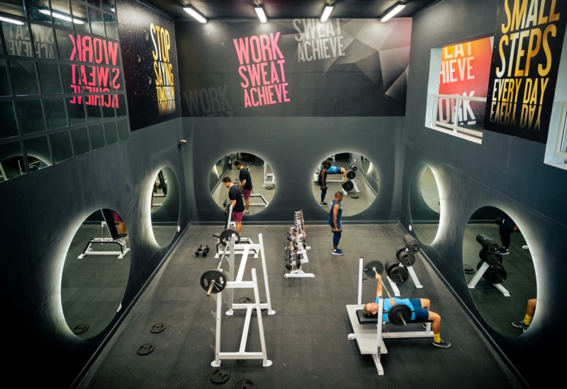 Hotel gym: dark studio with circular mirrors, motivational wall art, benches, free weights and guests lifting.