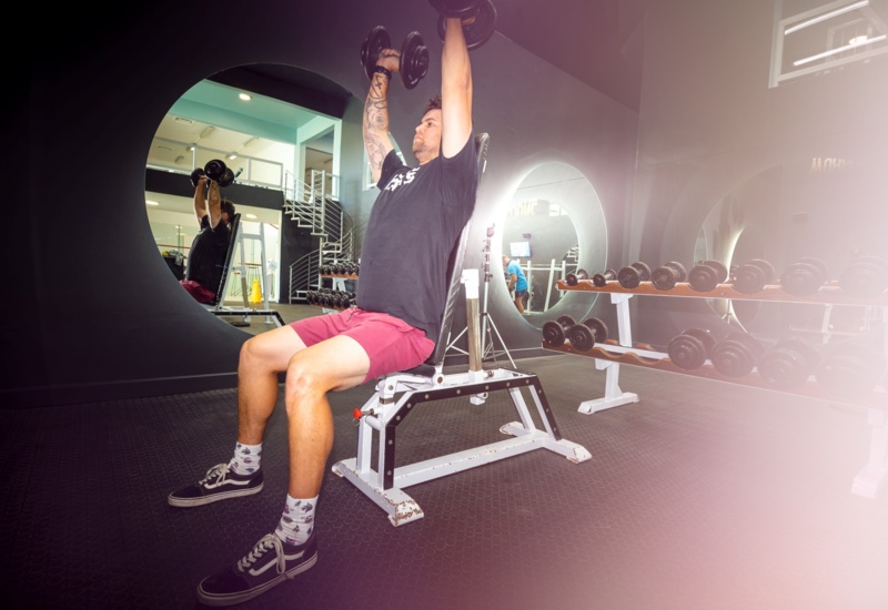 Guest doing seated dumbbell shoulder press on an adjustable bench in hotel gym with circular mirrors and dumbbell rack.