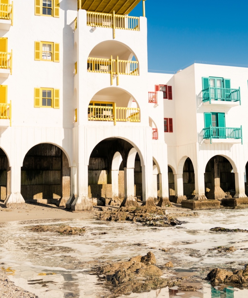 White beachfront hotel with colorful balconies and arched piers; mother and child stroll barefoot along the rocky shore.