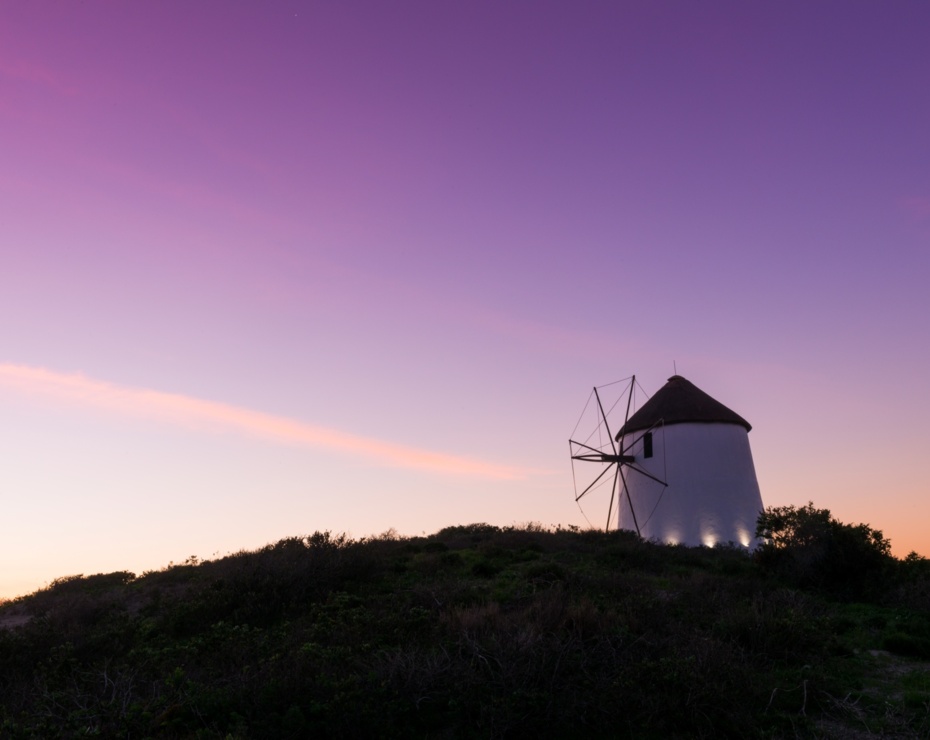 White hilltop windmill softly lit at dusk, framed by low scrub and a vivid purple-pink sunset sky — tranquil, romantic vista