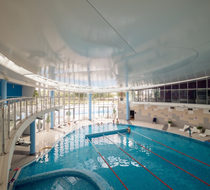 Geräumiges, modernes Hallenbad mit Schwimmbahnen und Ruhebereich an großen Fenstern mit Blick auf Grünflächen.