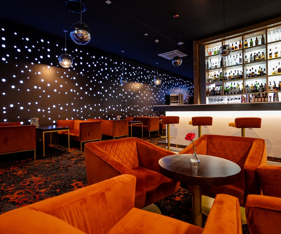 Nowoczesny hotelowy bar z wygodnymi pomarańczowymi fotelami, podświetlanym barem i dekoracyjną ścianą z białych punktów światła.