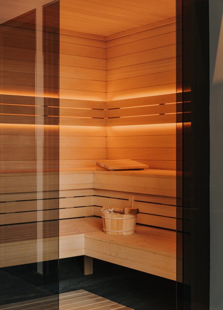 Nowoczesna sauna hotelowa wykończona jasnym drewnem, z ciepłym oświetleniem i drewnianym wiadrem z chochlą dla relaksu gości.