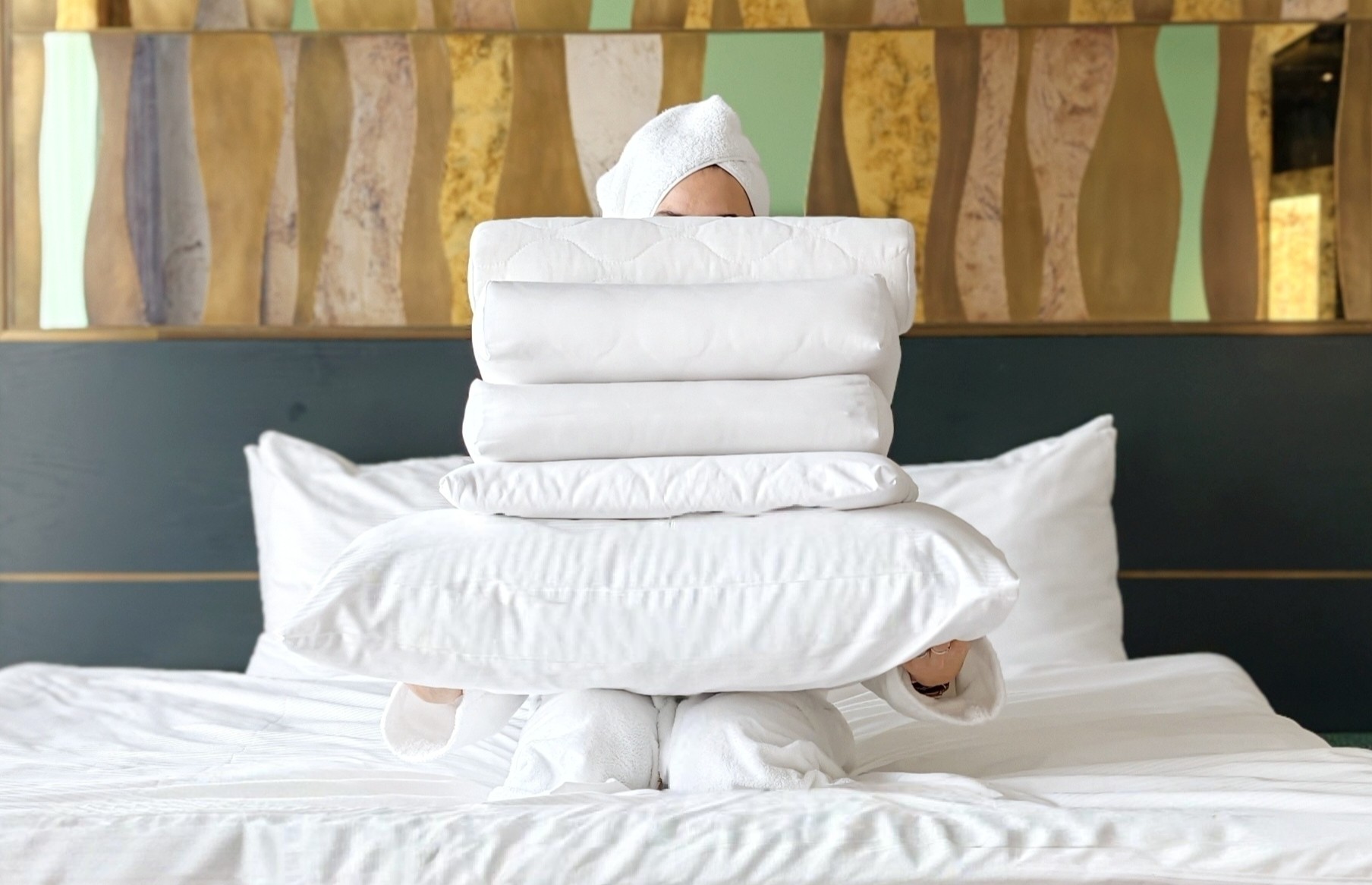 Gast im Zimmer des The Cyrus Hotels haelt frische weisse Bettdecken und Kissen auf einem Bett mit designorientiertem Kopfteil und kuenstlerischem Wandpanel, Atmosphaere von Sauberkeit, Komfort und Ruhe