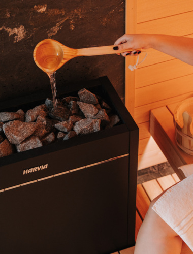 Hand giest Wasser mit einer Holzkelle über heiße Steine im Saunaofen umgeben von hellem Holz und entspannter Atmosphäre