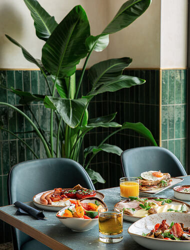 Tisch mit vorbereitetem Frühstück im Hotelrestaurant mit Tellern mit Brot, Eiern, Würstchen und frischen Beilagen, Gläsern mit Orangen- und Apfelsaft, vor grünen Wandfliesen und einer großen grünen Pflanze.