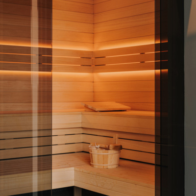 Nowoczesna sauna hotelowa wykończona jasnym drewnem, z ciepłym oświetleniem i drewnianym wiadrem z chochlą dla relaksu gości.