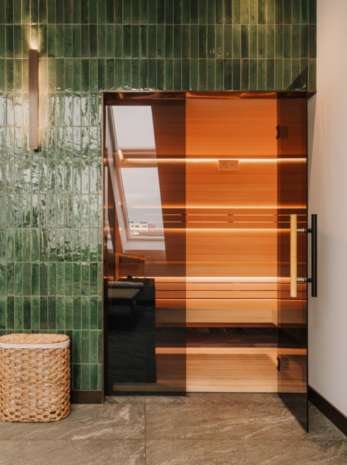 Nowoczesna sauna z drewnianym wnętrzem, szklanymi drzwiami i zieloną, płytkową ścianą w strefie wellness hotelu
