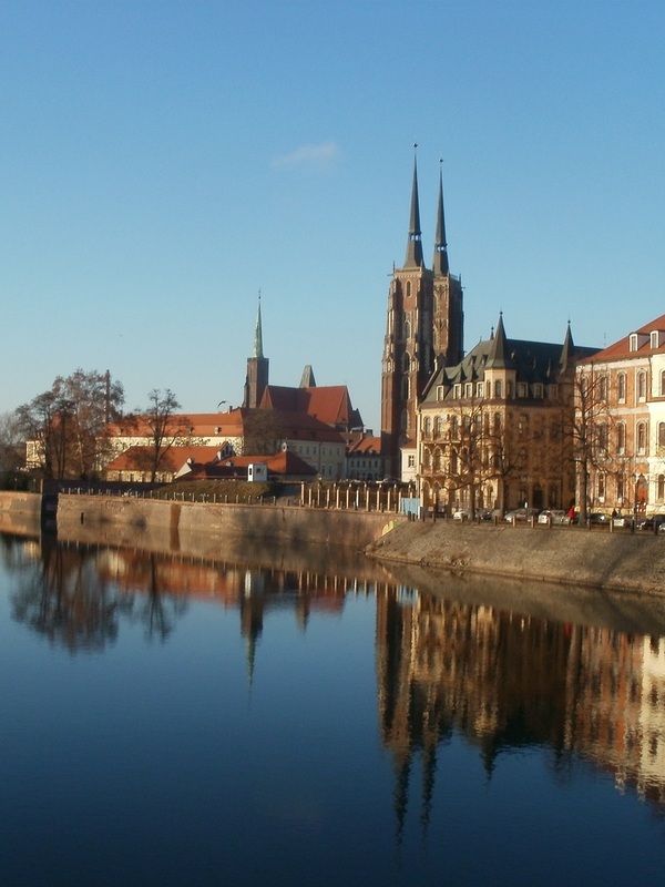Panorama der Dominsel in Breslau mit der gotischen Kathedrale St. Johannes des Täufers mit ihren zwei hohen Türmen, historischen Bürgerhäusern und dem Ufer, das sich an einem klaren Tag im ruhigen Wasser der Oder spiegelt.