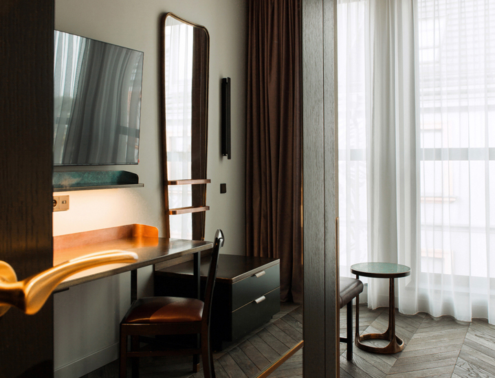 Nowoczesny hotelowy pokój z drewnianą podłogą, biurkiem, dużym lustrem oraz telewizorem na ścianie.