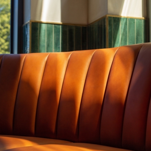 Brązowa, skórzana kanapa z pionowymi przeszyciami w przytulnym, dobrze oświetlonym wnętrzu z zielonymi płytkami.