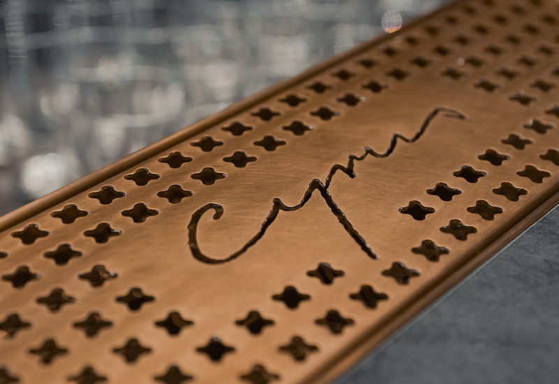 Miedziana barowa podkładka z ozdobnymi wycięciami i napisem, otoczona szklankami w tle