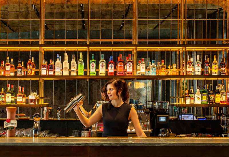 Barman przygotowująca koktajl za ladą w eleganckim barze z lustrzanymi ścianami i oświetleniem w ciepłych tonach.