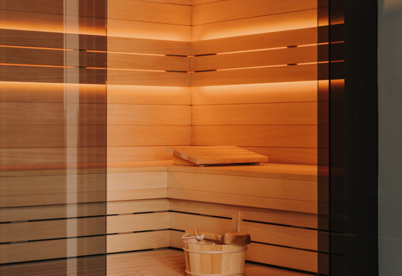 Nowoczesna sauna hotelowa z drewnianymi ławami, delikatnym podświetleniem i drewnianym wiadrem z chochlą.