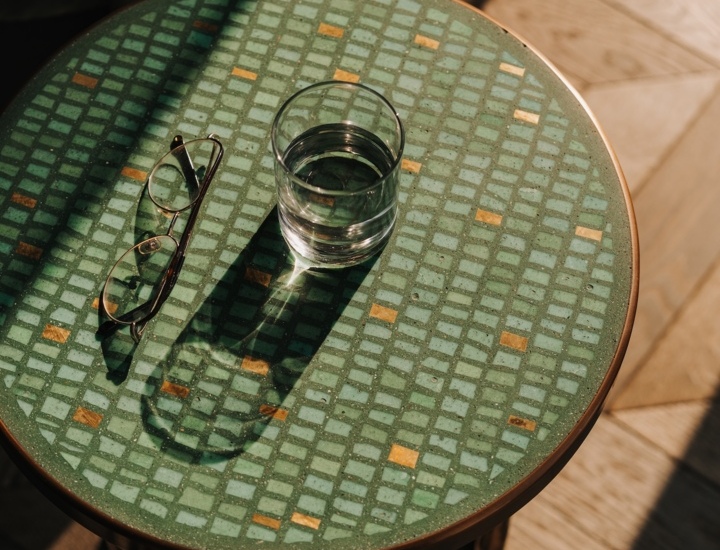Okrągły stolik z zielonym mozaikowym blatem, na którym stoi szklanka wody i leżą okulary, w słonecznym pokoju hotelowym.