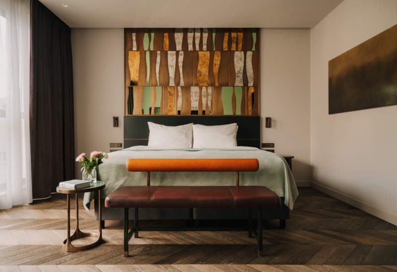 Nowoczesny pokój hotelowy z dużym łóżkiem, dekoracyjną ścianą nad zagłówkiem i elegancką ławką na drewnianej podłodze