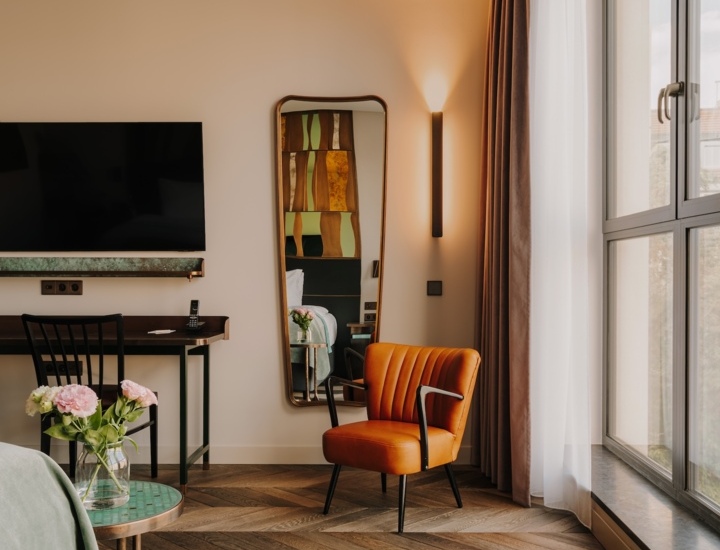 Nowoczesny pokój hotelowy z dużym oknem, pomarańczowym fotelem, biurkiem, lustrem i drewnianą podłogą.