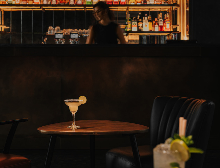 Elegancki bar hotelowy z półkami pełnymi alkoholi, ciemnymi meblami i dwoma kolorowymi drinkami na stołach.