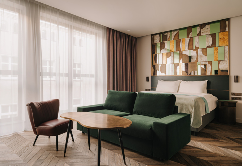 Nowoczesny pokój hotelowy z dużym łóżkiem, zieloną sofą, brązowym fotelem i dekoracyjną mozaiką na ścianie