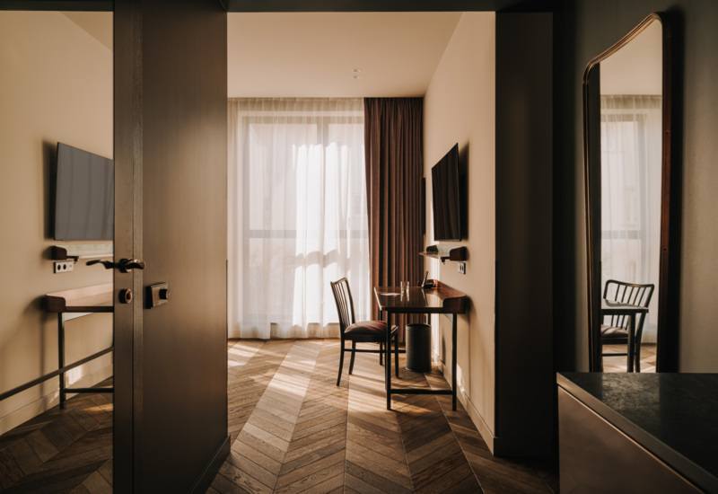 Przestronny pokój hotelowy z drewnianą podłogą, biurkiem, krzesłem i dużym lustrem, jasne zasłony wpuszczają światło