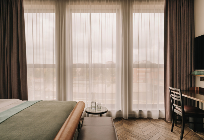 Przytulny pokój hotelowy z dużym łóżkiem, stylowymi zasłonami, drewnianą podłogą oraz stolikiem z karafką i szklanką wody.