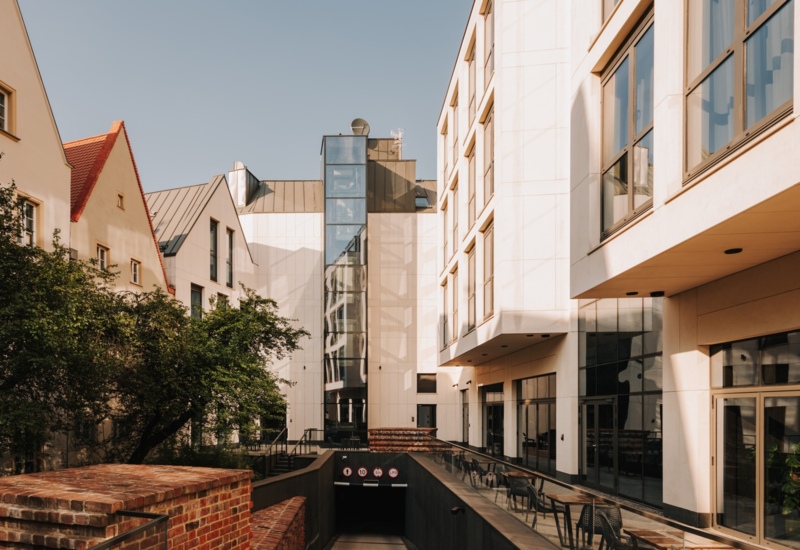 Nowoczesny hotel z białą elewacją, dużymi oknami, tarasem z miejscami do siedzenia i zielonym podwórkiem