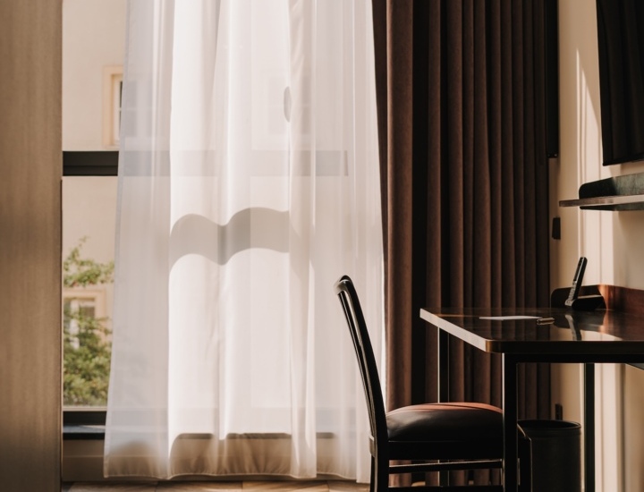 Pokój hotelowy z drewnianą podłogą, biurkiem, krzesłem i dużym oknem z zasłonami przepuszczającymi światło